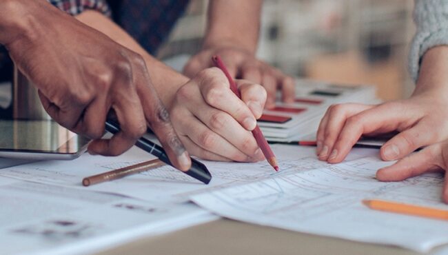 A close-up of several hands holding pens and pointing to a printed floor plan.