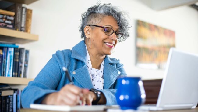 A person smiling at a laptop screen and taking notes in a notebook on their desk.